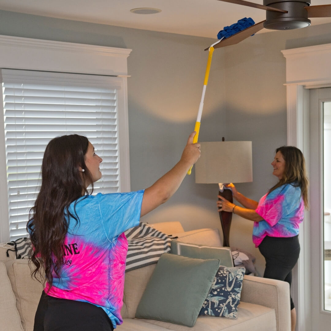 Two women cleaning a bright living room; one uses an extendable duster to clean a ceiling fan while the other wipes down a lamp base.
